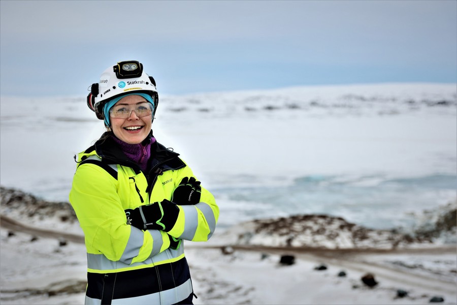 Uma mulher vestida com EPIs e uniforme da Statkraft está de braços cruzados e sorri em uma paisagem com neve.
